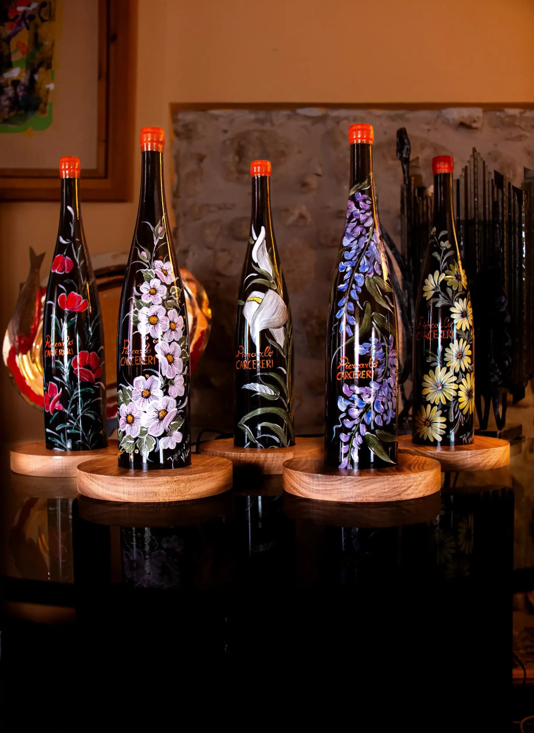 Five dark wine bottles from the 'Piercarlo CARCERERI' series, each hand-painted with a unique floral design, standing on wooden bases on a dark reflective surface indoors.