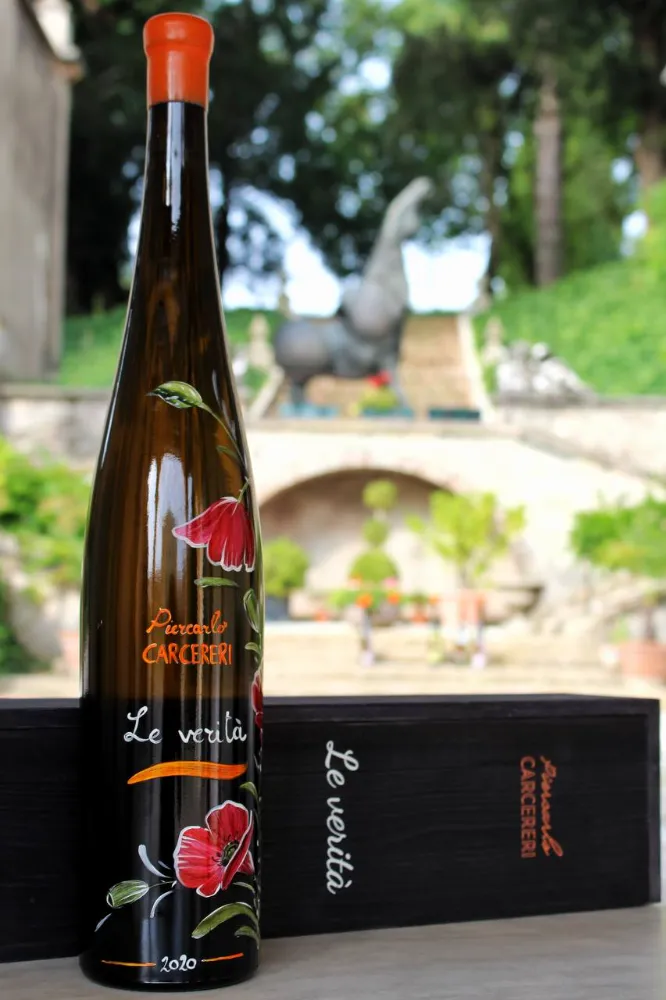 Close-up of a dark wine bottle hand-painted with red poppies, labeled 'Piercarlo CARCERERI Le verità 2020', next to a black wooden box in a garden setting.