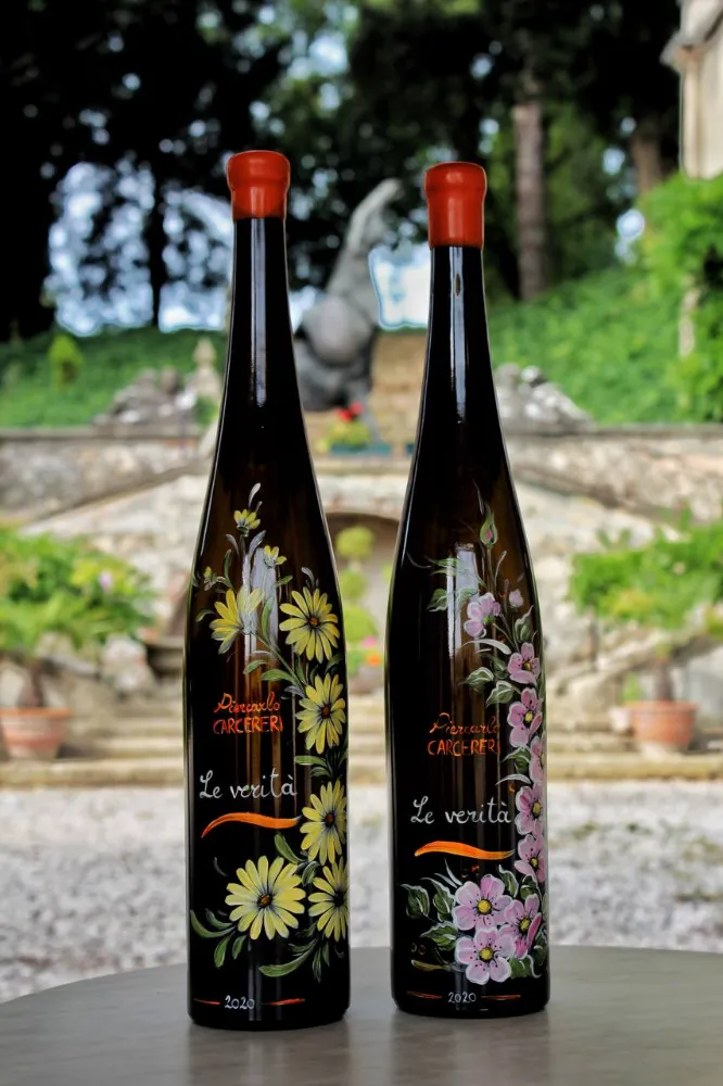 Two dark wine bottles from the 'Le verità 2020' series, hand-painted with yellow daisies and pink cherry blossoms, set against a stone staircase backdrop.